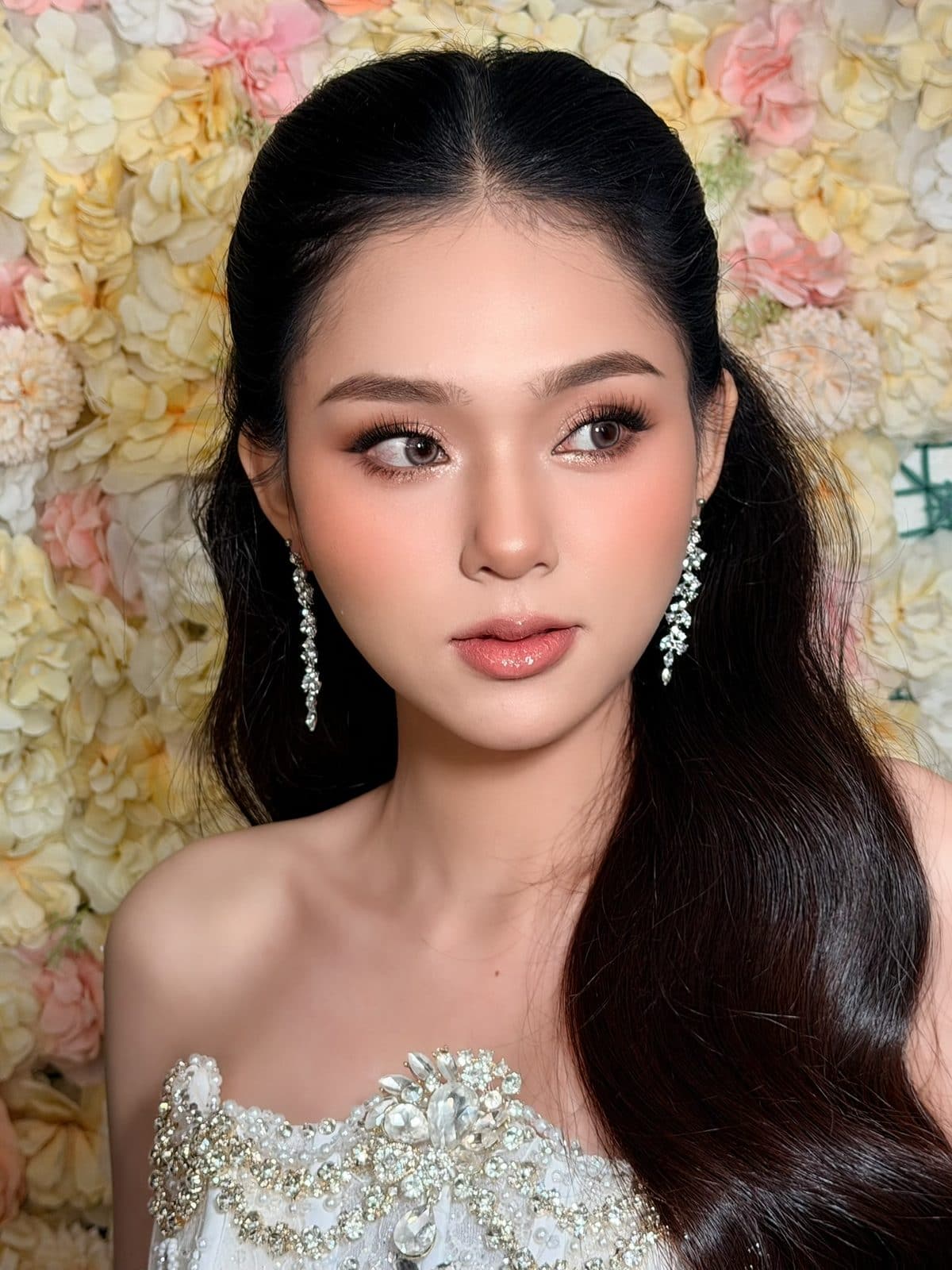 Cô dâu buộc tóc nửa đầu, bông tai đá dài, váy cưới đính đá trước phông hoa pastel, KINKAY Sài Gòn